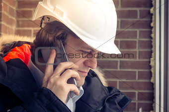 Construction inspector in white helmet talking on mobile phone on the brown brick wall background. Soft focus, toned. Close up.