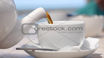 White teapot pouring tea into cup in beach cafe by the sea with sunlight.