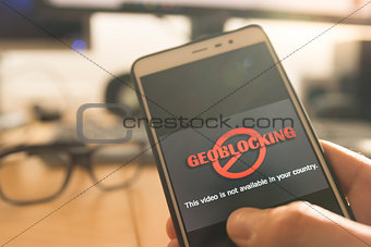 Hands using a smartphone with Geo-blocking on screen.