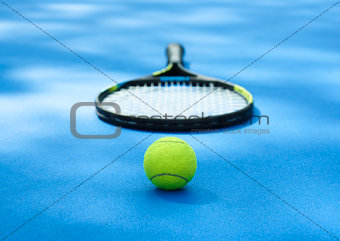 Tennis ball is laying near racket on blue cort carpet.