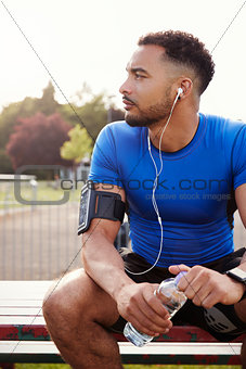 Young male athlete sitting on park bench holding water bottle