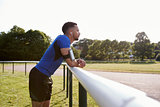 Male athlete at track leaning on fence, three quarter length