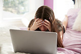 Upset teen girl lying on bed with head in hands using laptop