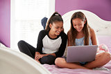 Two teen girls sitting on bed using a laptop, close up