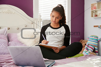 Teen girl sitting on bed watching computer and writing notes