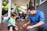 Father And Son Sit On Porch Of House Playing With Toys Together