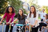 Three pre-teen girls on scooters and a bike at a block party