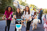 Three pre-teen girls on scooters and a bike at a block party