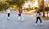 Three girls riding on scooters and a bike in the street