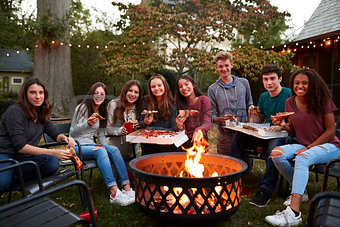 Teenagers at a fire pit with take-away pizzas look to camera