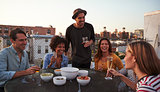 Six adult friends laughing at a table on a rooftop, close up