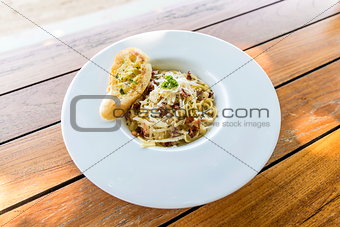 The spaghetti carbonara with cheese ,bacon and crackers on top. In a white plate on a brown wooden table.