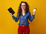 happy woman isolated on yellow showing coffee cup and tablet PC