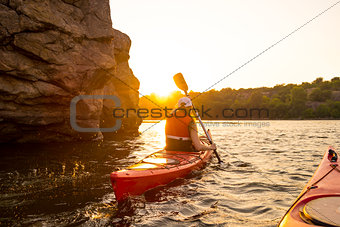 Young Woman Paddling the Red Kayak on Beautiful River or Lake near High Rocks at Sunset