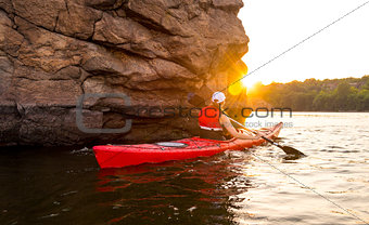 Young Woman Paddling the Red Kayak on Beautiful River or Lake near High Rocks at Sunset