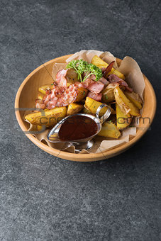 Fried potatoes with appetizing bacon and sauce are in a wooden plate