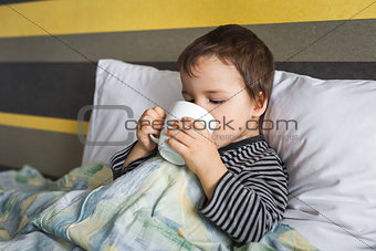 sad ill curing baby boy sitting on bed drinking medicament