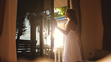 Pretty girl opens the door in the early morning during sunrise with lens flare effects and go out to the terrace