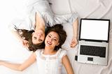 Two Young girls relax and smile with laptop lying on the floor
